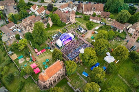 Aerial view of a festival in a historic setting with stage and many visitors surrounded by trees,