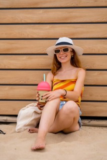 Young woman wearing a hat and sunglasses, smiling while holding a colorful drink on a sunny day at
