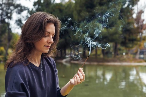 A young Caucasian woman with long brown hair sits by a pond, practicing mindfulness. She holds a