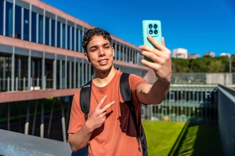 Young male student smiling while capturing a selfie with his smartphone, showing a peace sign on a