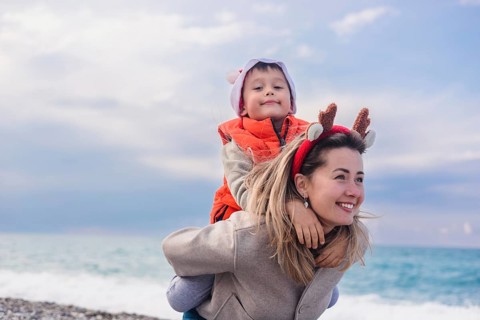 A young Caucasian woman with long blonde hair carries a smiling Asian boy on her back at the beach