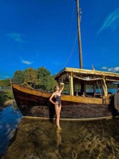 Wide angle view of a slender woman in a black swimsuit standing in a lake near an old wooden ship