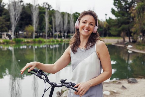 A young Caucasian woman with brown hair stands beside a bicycle near a serene lake. She wears pink