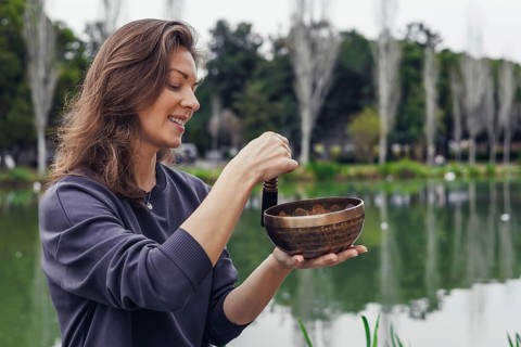 A young Caucasian woman with long brown hair holds a wooden bowl near a serene lake. Trees and