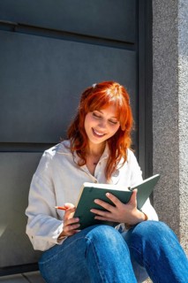 Young redhead woman sitting outside, smiling while writing notes in a green notebook, enjoying a