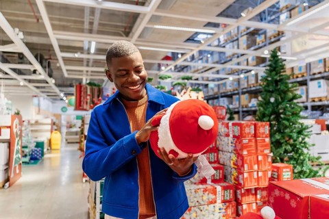 A young Black man smiles while holding a large red Santa toy in a festive store filled with