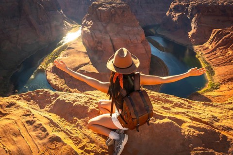 Young woman hiker sitting on a sandstone cliff, arms outstretched, feeling freedom and happiness