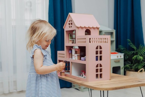 Focused child in a blue dress plays with a dollhouse on a wooden table inside a room