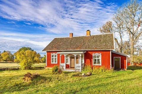 Red wooden cottage with white trim stands in a rural landscape surrounded by autumn trees. Bright