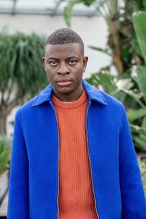 A young African man stands in a lush green environment. He has short black hair and wears a blue