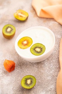 Yogurt with kiwi in white bowl on brown concrete background and orange linen textile, side view,
