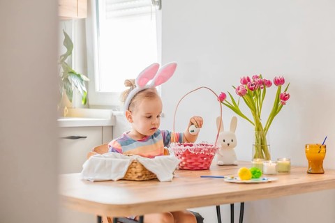 A young child wears bunny ears and dips an Easter egg into a basket at a table. Flowers and candles