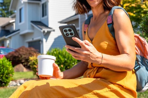 Woman in a yellow dress and backpack interacting with a smartphone while holding a coffee cup.