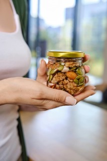 Person stands indoors in bright room with large windows holding a jar of honey filled with mixed