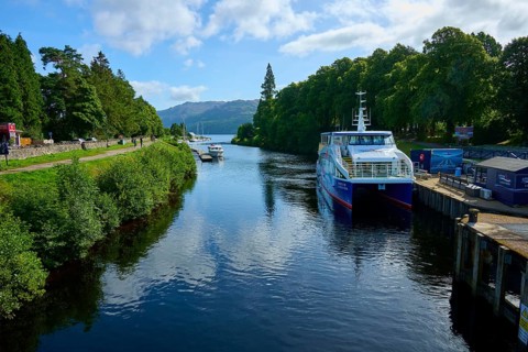 A large boat sails on a quiet river surrounded by trees and rolling hills, Caledonian Canal, Fort