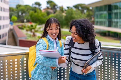Two smiling multi-ethnic female students having fun while reading notes in the university campus