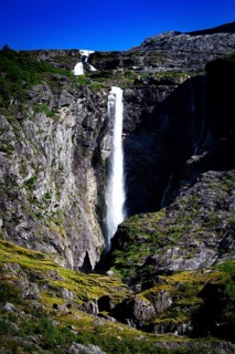 Tall waterfall plunging into a rocky gorge surrounded by green vegetation and blue sky, view of a