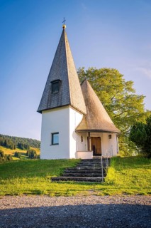 White chapel with pointed roof on green meadow, surrounded by trees, under blue sky, Bruder Klaus