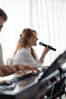 A young Caucasian woman with curly hair sings into a microphone. A hand plays a keyboard in the