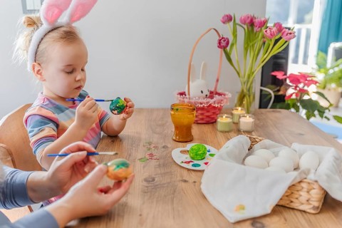 Little caucasian girl in bunny ears painting Easter eggs with mother at home. Spring holiday