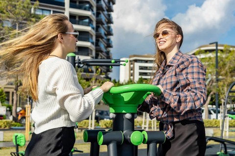 Two young women engage in outdoor exercise at a fitness station in an urban park. One has long