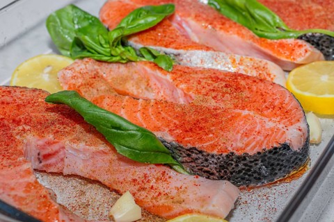 Fresh salmon fillets arranged in a glass dish with lemon slices and basil leaves. The fish is raw