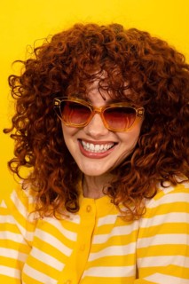 A woman with vibrant curly hair smiles confidently, wearing fashionable sunglasses and a