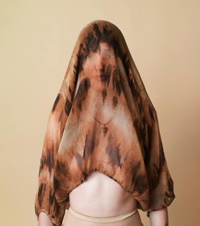 A young woman stands against a beige studio background, covering her head with a sheer, patterned