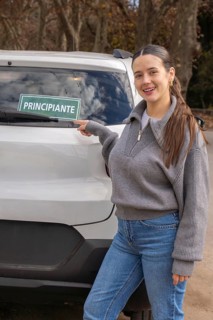 Smiling young woman pointing at principiante beginner driver sign on rear car window, autumn
