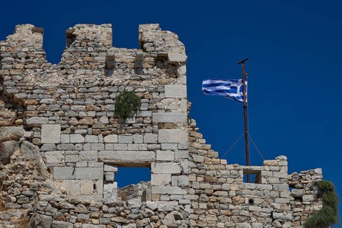 Old stone ruins with a waving Greek flag against a deep blue sky, Johanniter fortress from the 14th