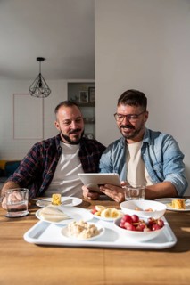 A joyful gay couple sits at a cozy breakfast table, engaged with a digital tablet. Their warm