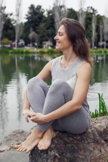 A young Caucasian woman with long brown hair sits on a rock by a calm lake. She wears a light gray