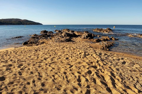 Beach-level viewpoint toward a rocky outcrop on sandy Gigaro Beach with footprints, calm sea,