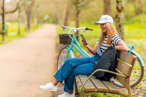 Cheerful young woman using smartphone while sitting on a bench in a park with her bicycle next to
