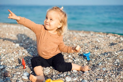 A joyful child sits on a sunny beach, happily playing with sand and pebbles. The clear blue sea