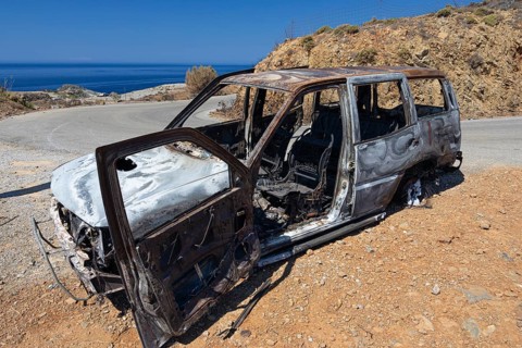 A rusty, burnt out car with a view of the coastal sea and dry hills, forest fire, fire damage, fire