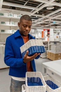 A young Black man examines a blue and white storage basket in a modern retail store. Shelves filled