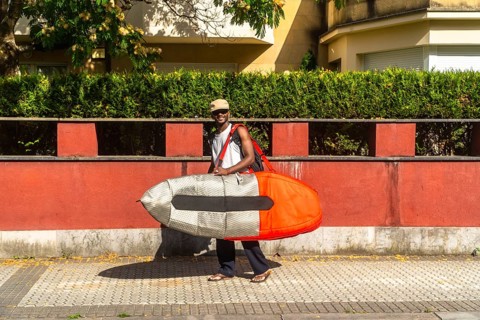 Happy young man walking along a sunny urban street carrying a surfboard in a protective bag, ready