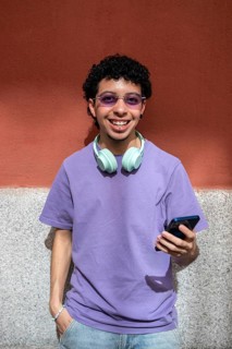 A LGBTQ+ cheerful person stands against a wall, wearing headphones and holding a phone. They exude