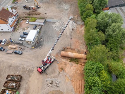 Aerial view of a construction site with a crane moving material, surrounded by trees and houses,
