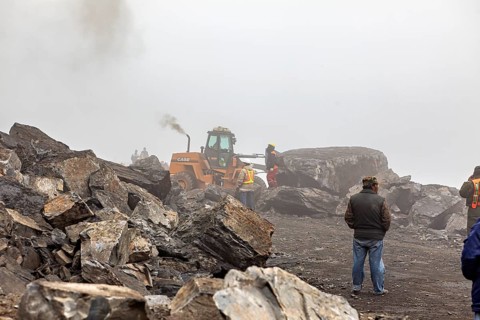 Excavator picks up large rocks on foggy construction site, workers nearby, rock slide on the pass