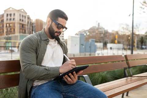 A man wearing sunglasses sits on a park bench, deeply focused on sketching on a digital tablet with