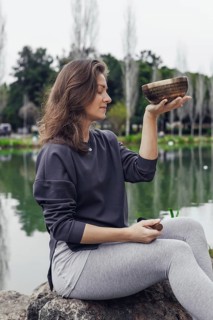 A young Caucasian woman with brown hair sits by a lake, holding a bowl. She wears a gray sweater