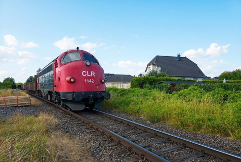 Red train on rails against green background with house in summer under cloudy sky, NoHab diesel