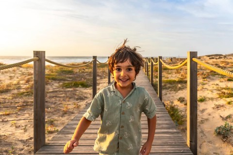 Child running towards the camera on a wooden boardwalk, smiling widely, with a sandy beach and
