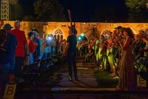Artist walks with raised guitar through applauding audience at night concert, Calwer Klostersommer