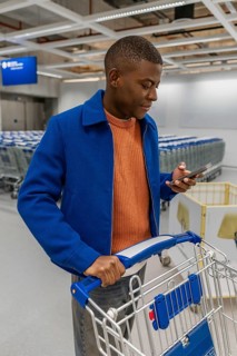 A man stands in a store and looks at his phone while holding a shopping cart. He wears a blue