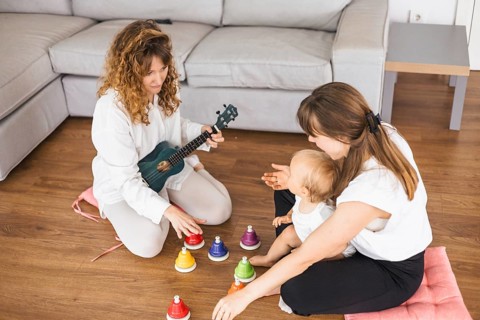 Woman playing ukulele with a baby close by, surrounded by colorful bells indoors