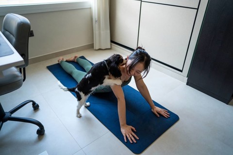 A young Asian woman attempts to exercise on a yoga mat at home, while her playful dog eagerly