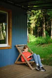 Young woman sits on wooden chair on terrace of tiny house and reading book. Trees and greenery